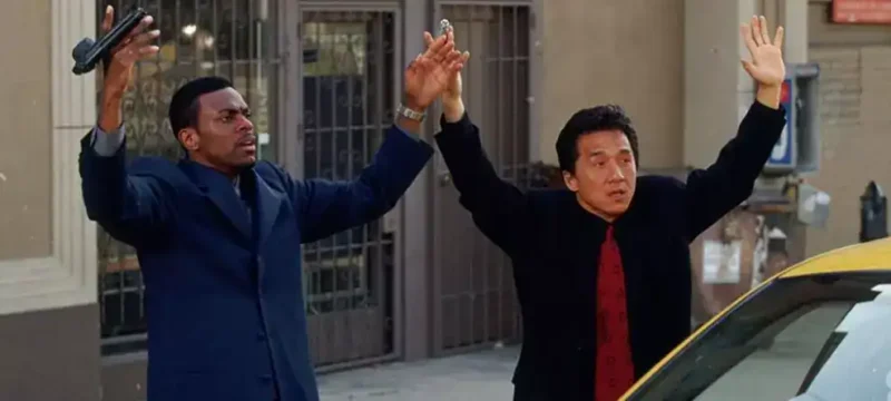 Cena de A Hora do Rush 4: dois homens em ternos escuros levantam os braços em uma rua movimentada, ao lado de um táxi amarelo. Um deles segura uma arma, enquanto o outro parece rendido. O clima mistura tensão e humor, marca registrada da franquia estrelada por Jackie Chan e Chris Tucker.