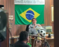 Ator Jim Caviezel caracterizado para interpretar Jair Bolsonaro no filme "Dark Horse", sentado em escritório com Bandeira do Brasil e a frase "Brasil Acima de Tudo" ao fundo.